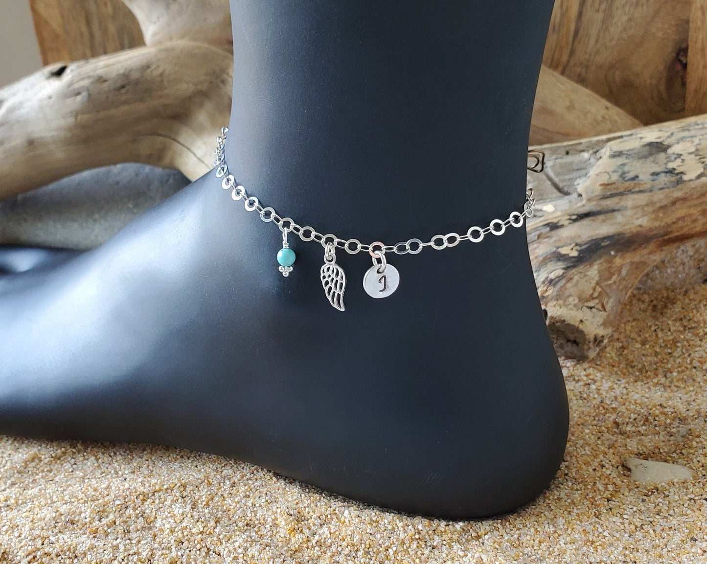 Deluxe Personalized Wing, Initial, Birthstone Anklet, Ankle Bracelet, pendant dangling on Silver chain on black display on sand 