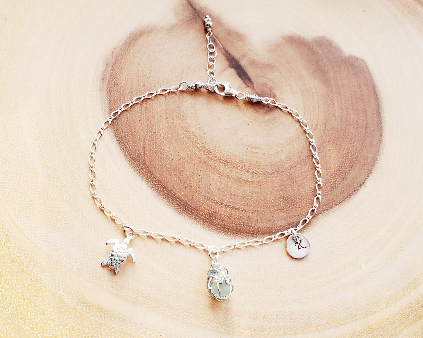 Sterling Silver Aqua Blue Beach Glass/ Sea Glass Anklet, Ankle Bracelet with a Turtle, round Initial pendant and Celtic Eternity Coil pendant on flat figaro chain on wood background 