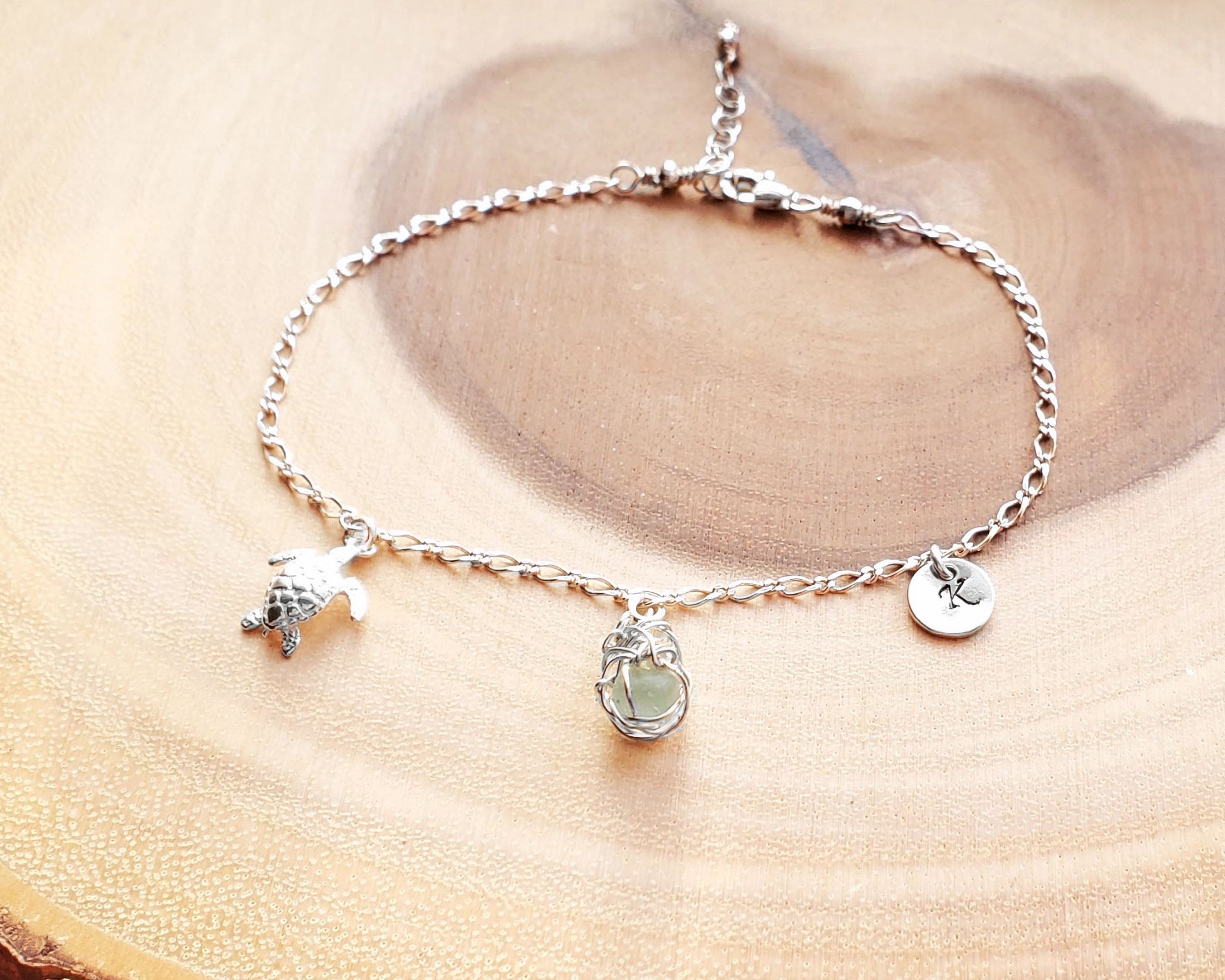 Sterling Silver Aqua Blue Beach Glass/ Sea Glass Anklet, Ankle Bracelet with a Turtle, round Initial pendant and Celtic Eternity Coil pendant on flat figaro chain on wood background 
