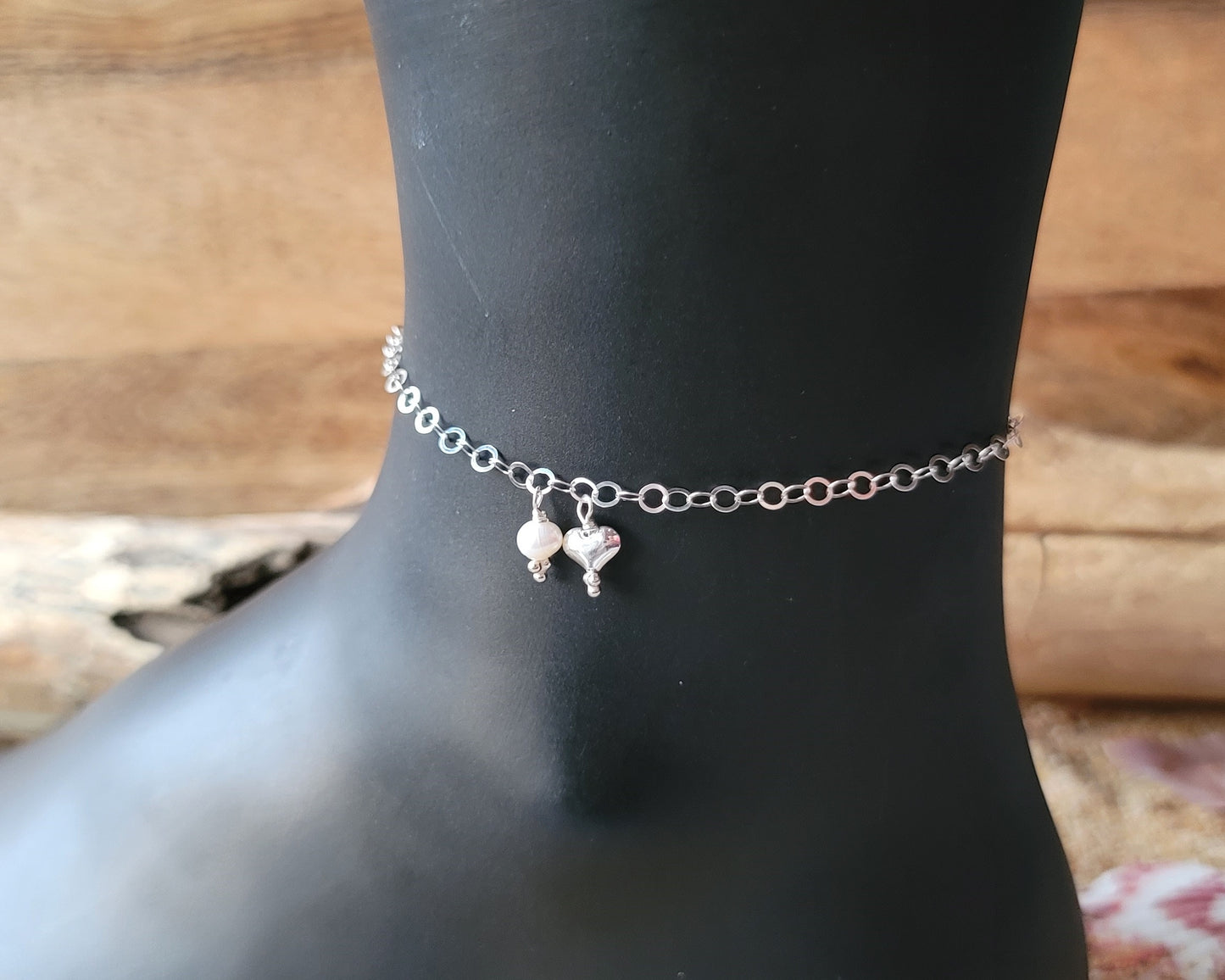 Silver Chain Anklet Ankle Bracelet with a small dangling heart and pearl on black foot with wood background 
