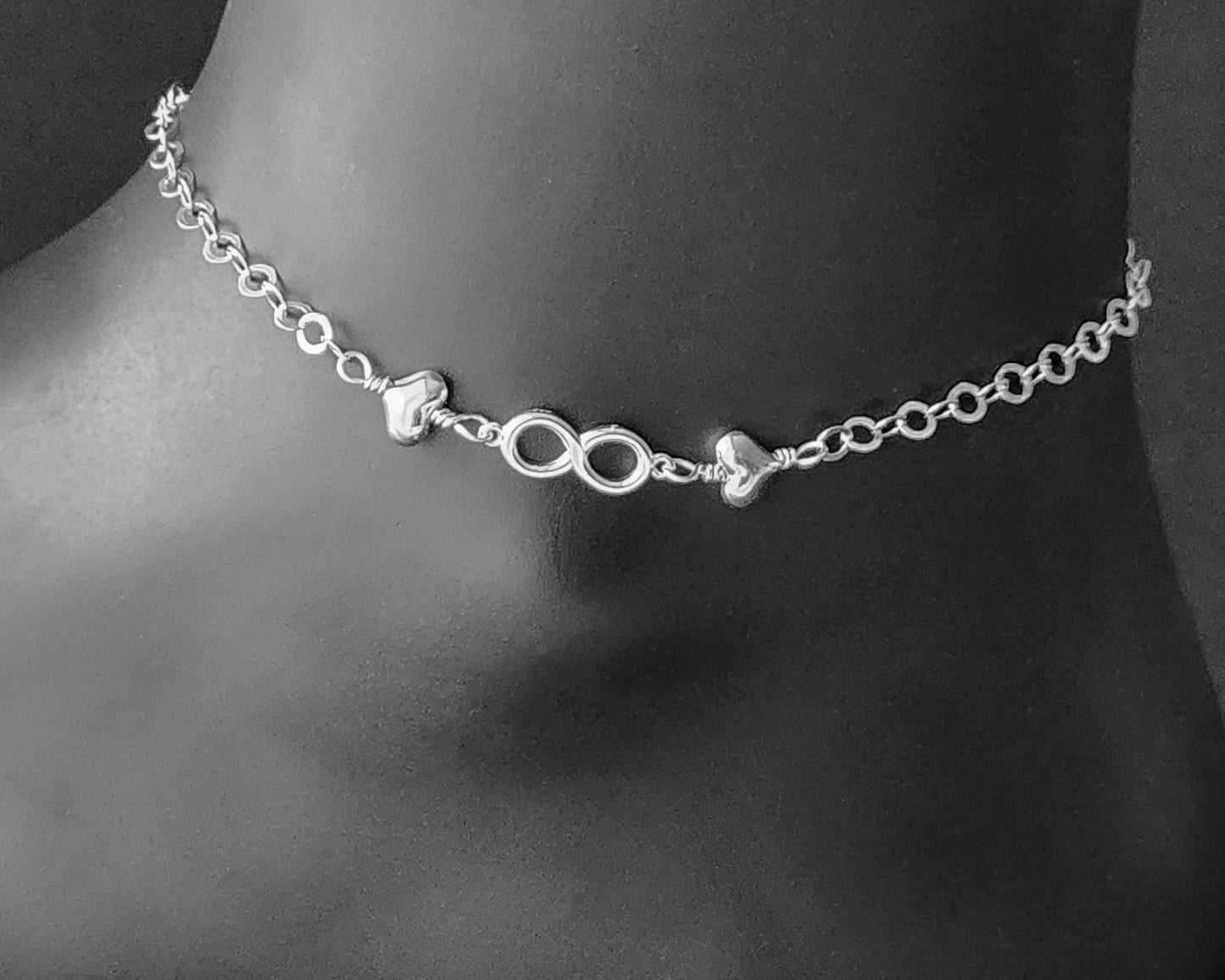 Silver Anklet with infinity pendant and two hearts on either side on chain displayed on black ankle