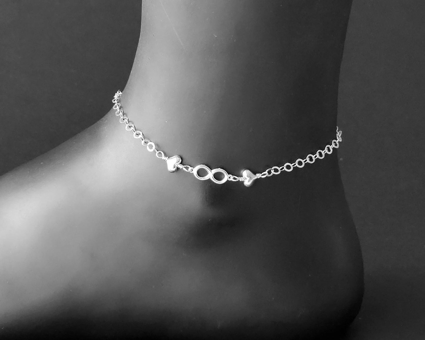 Silver Anklet with infinity pendant and two hearts on either side on chain displayed on black ankle