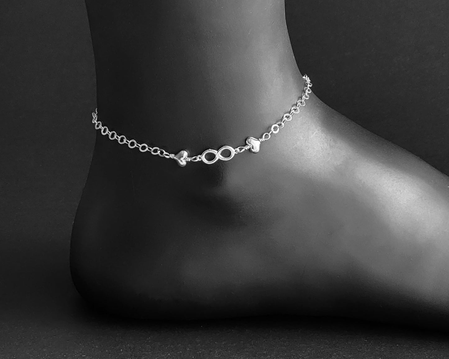 Silver Anklet with infinity pendant and two hearts on either side on chain displayed on black ankle