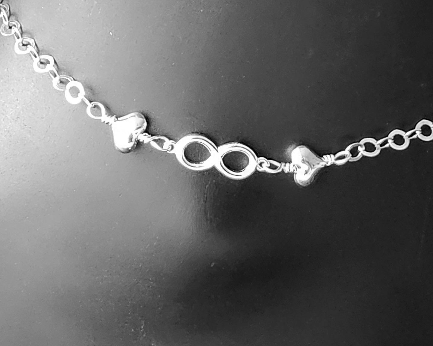 Silver Anklet with infinity pendant and two hearts on either side on chain displayed on black ankle