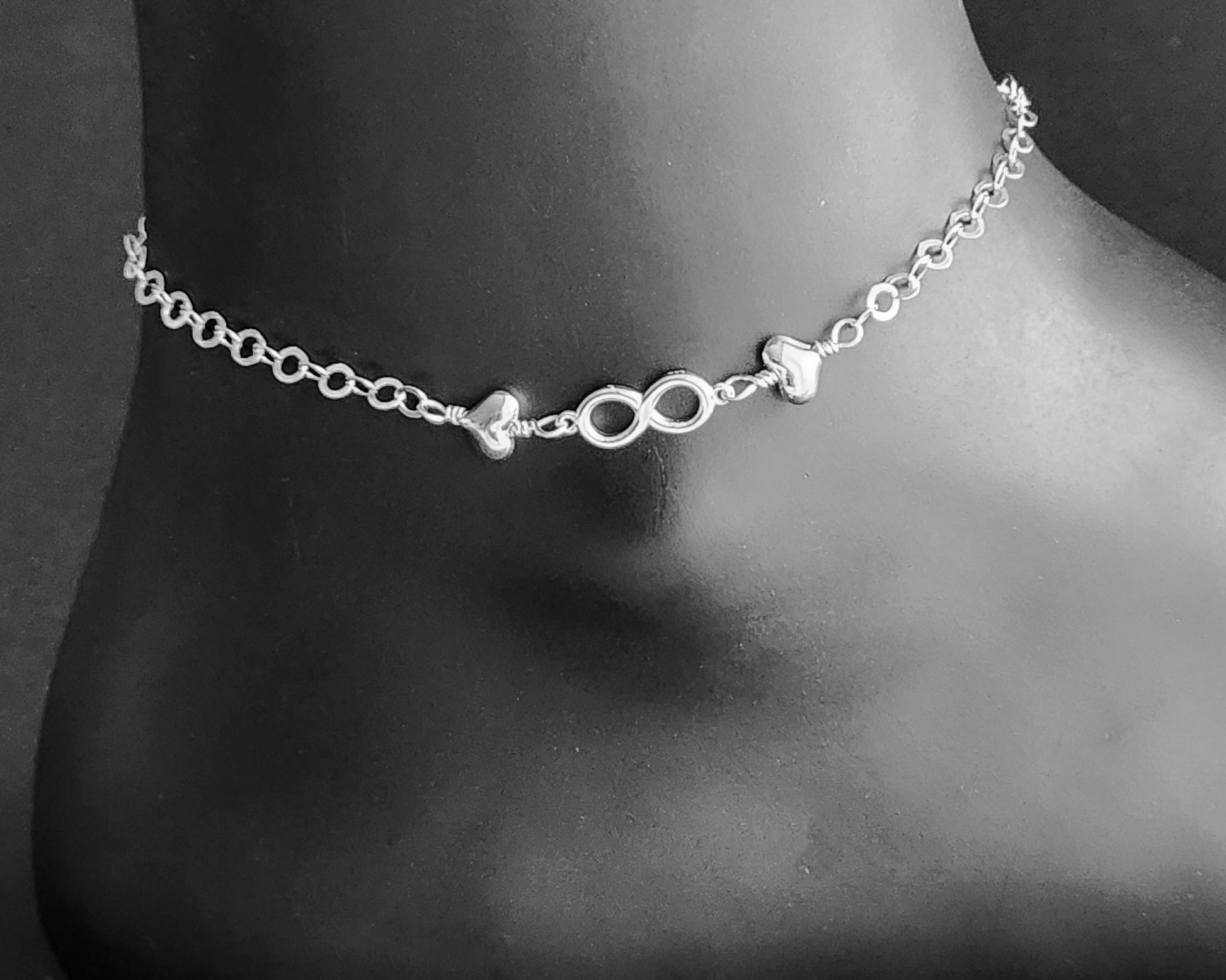 Silver Anklet with infinity pendant and two hearts on either side on chain displayed on black ankle
