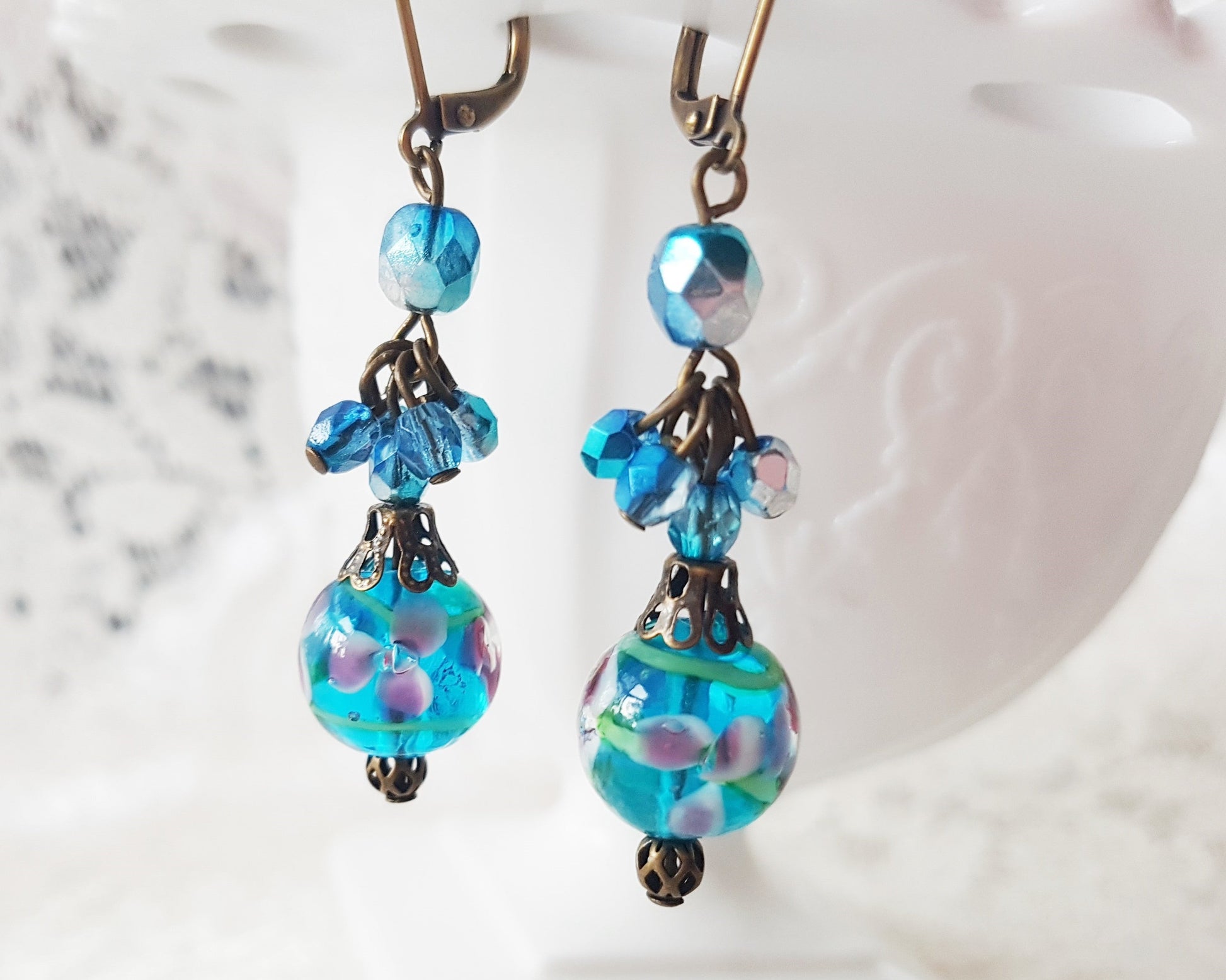 Long Blue Floral Vintage Inspired Earrings made with deep aqua blue beads, floral beads, and sparkly clusters above. The earring dangle on white vase on white lace
