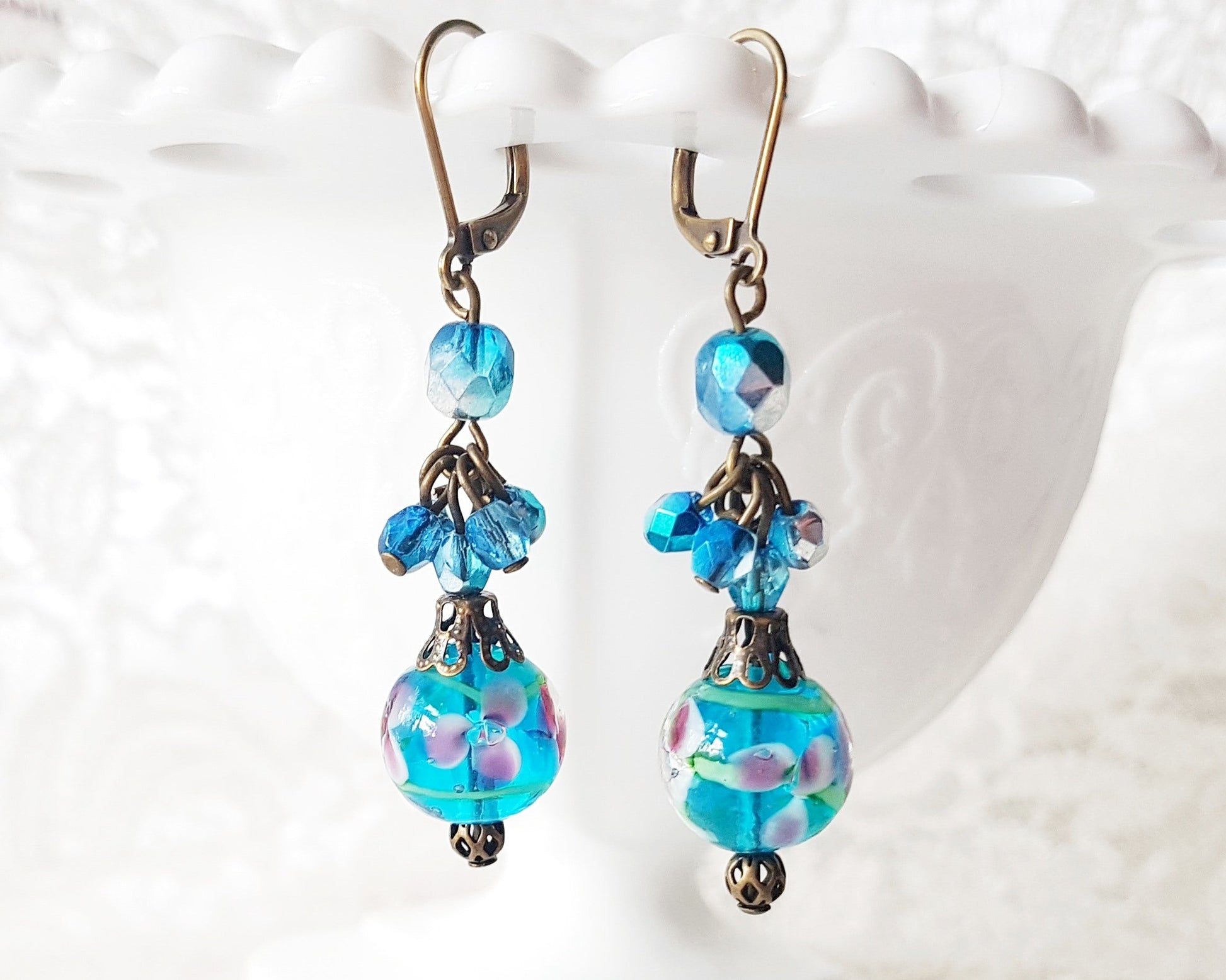 Long Blue Floral Vintage Inspired Earrings made with deep aqua blue beads, floral beads, and sparkly clusters above. The earring dangle on white vase on white lace