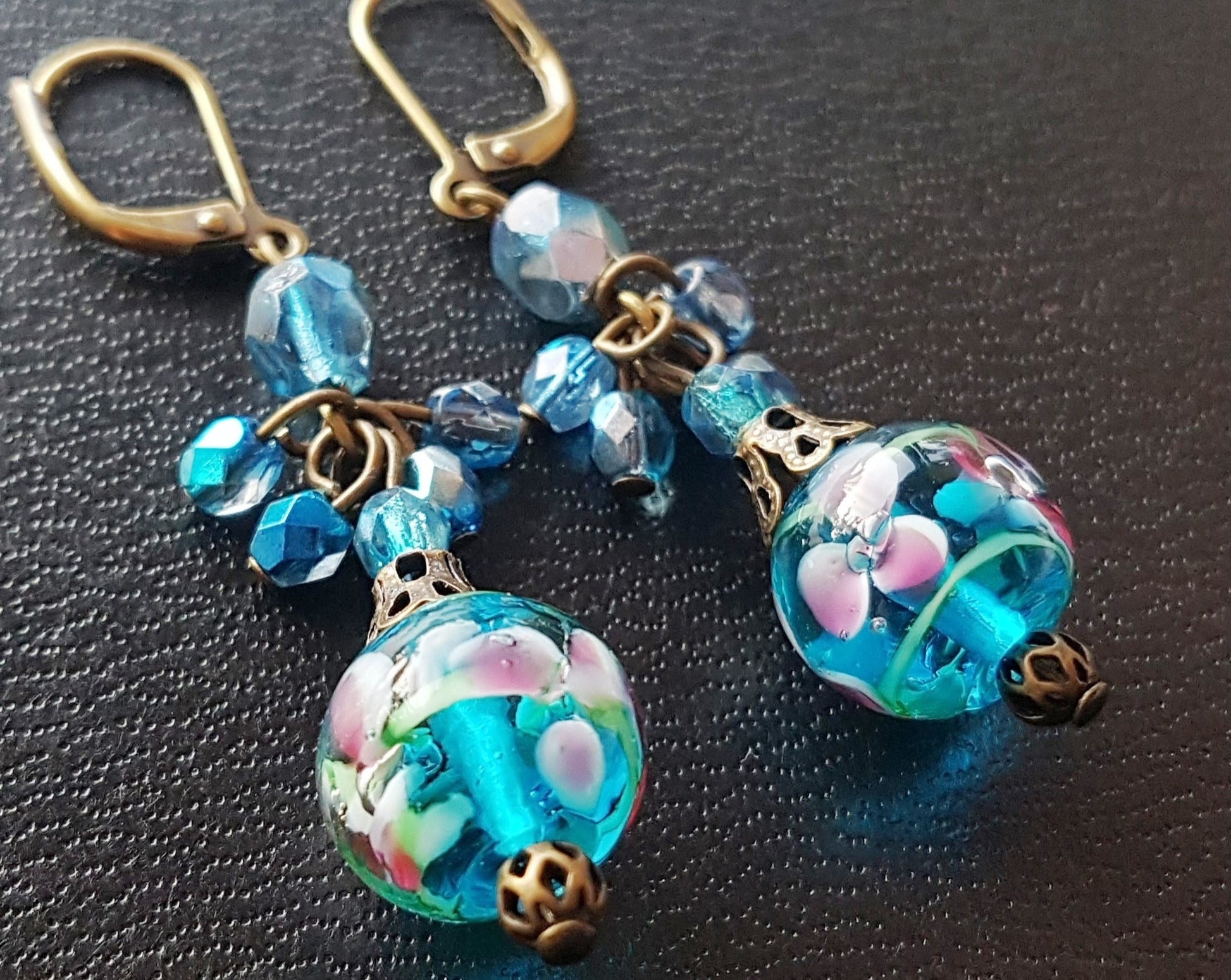 Long Blue Floral Vintage Inspired Earrings made with deep aqua blue beads, floral beads, and sparkly clusters above. The earrings are displayed on Black surface