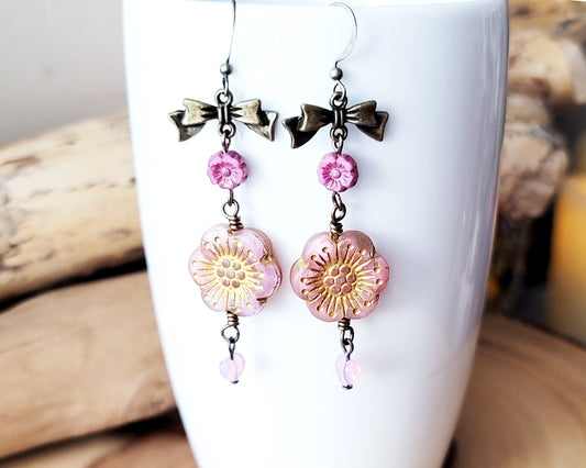 Long Vintage Romance Pink Flower Bow Earrings, with little pink heart dangles on the base, displayed on white cup