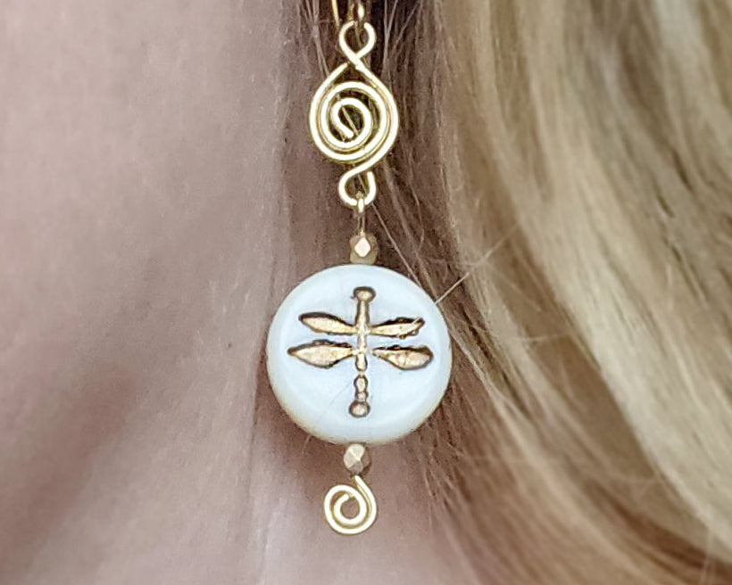 Gold and white dragonfly earrings with Celtic Coils and french style earring hooks worn by a person with blonde hair.
