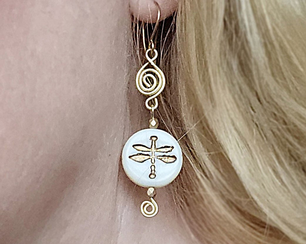 Gold and white dragonfly earrings with Celtic Coils and french style earring hooks worn by a person with blonde hair.