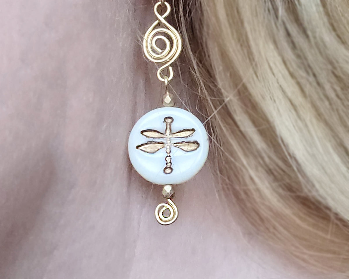 Gold and white dragonfly earrings with Celtic Coils and french style earring hooks worn by a person with blonde hair.