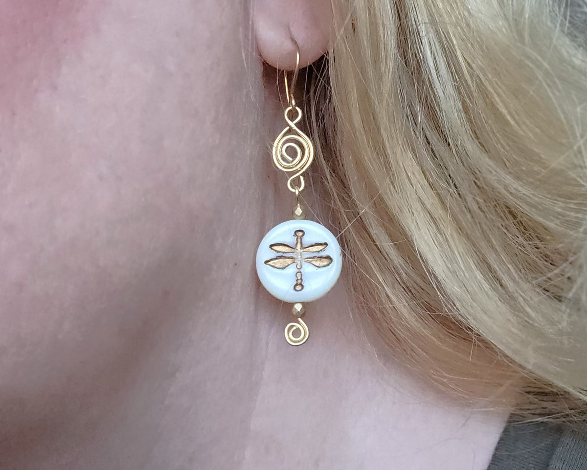 Gold and white dragonfly earrings with Celtic Coils and french style earring hooks worn by a person with blonde hair.