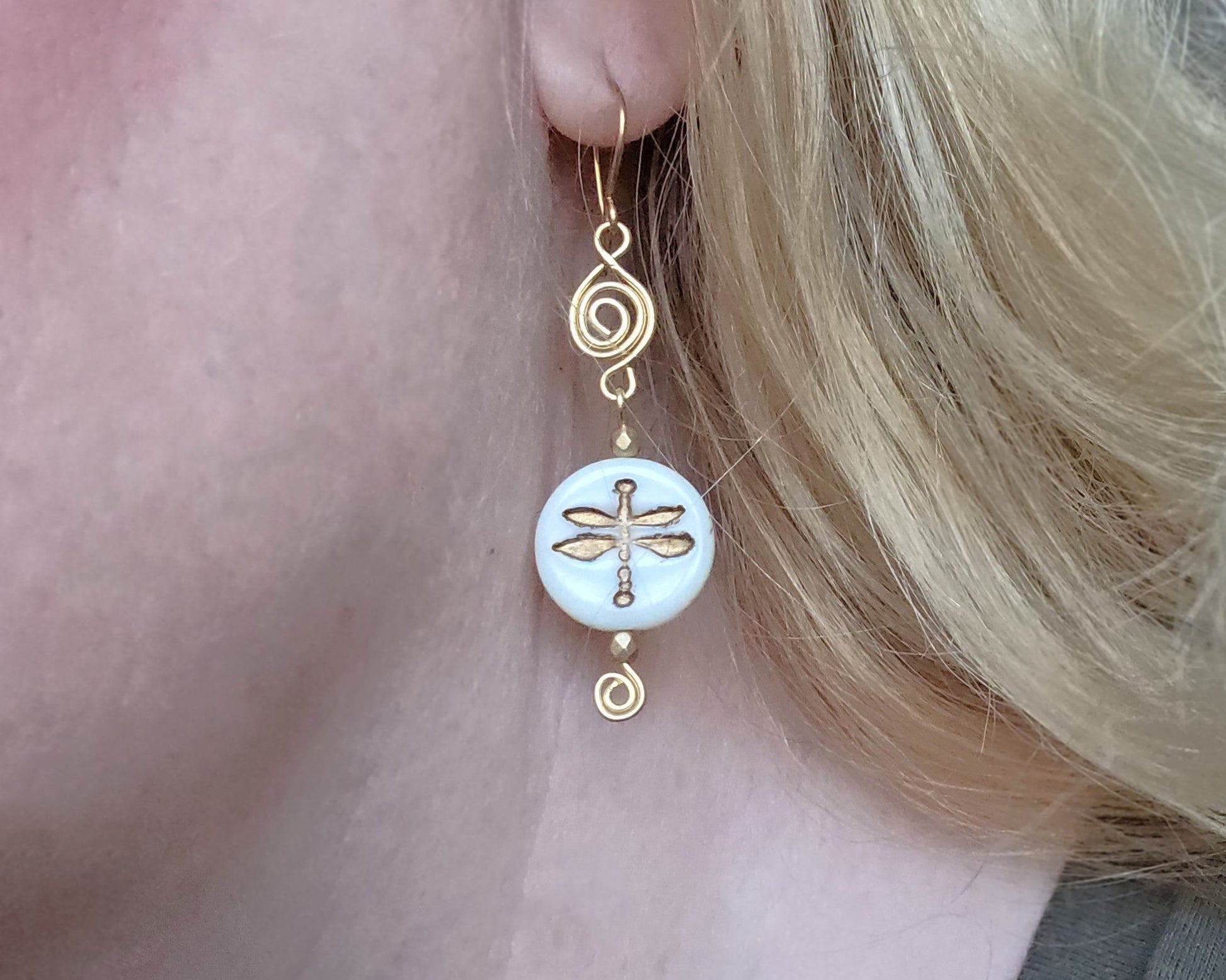 Gold and white dragonfly earrings with Celtic Coils and french style earring hooks worn by a person with blonde hair.