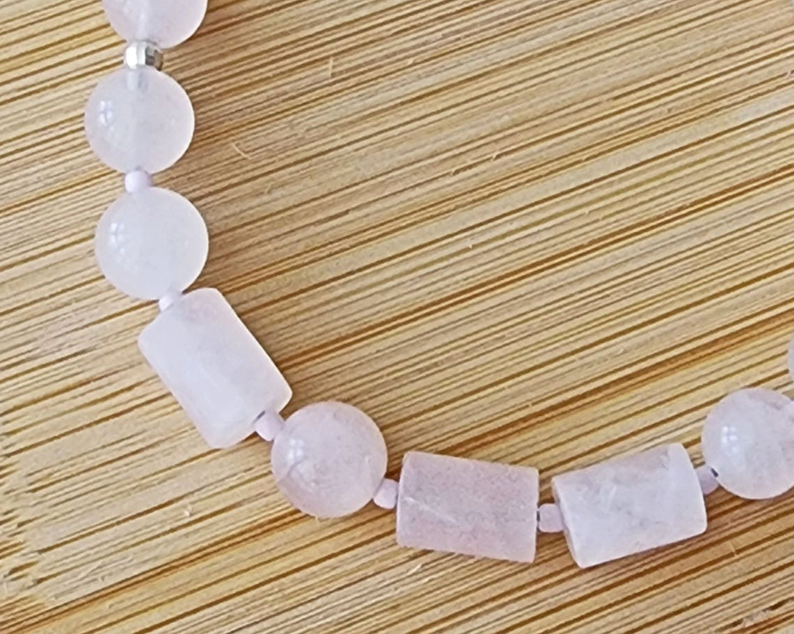 Rose Quartz PEACE bracelet, Morse Code spells the word PEACE with round and tube shaped pale pink Rose Quartz beads displayed on wood.