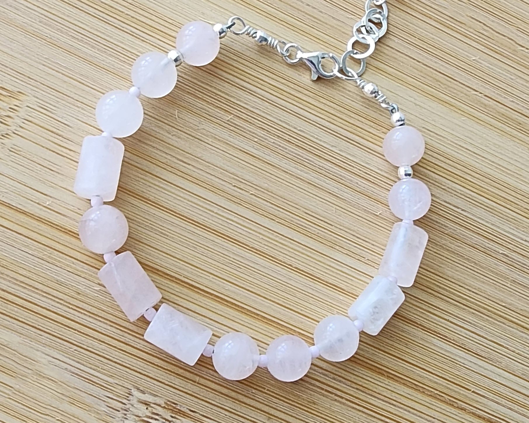 Rose Quartz PEACE bracelet, Morse Code spells the word PEACE with round and tube shaped pale pink Rose Quartz beads with Sterling Silver lobster claw clasp and extension chain, displayed on wood.