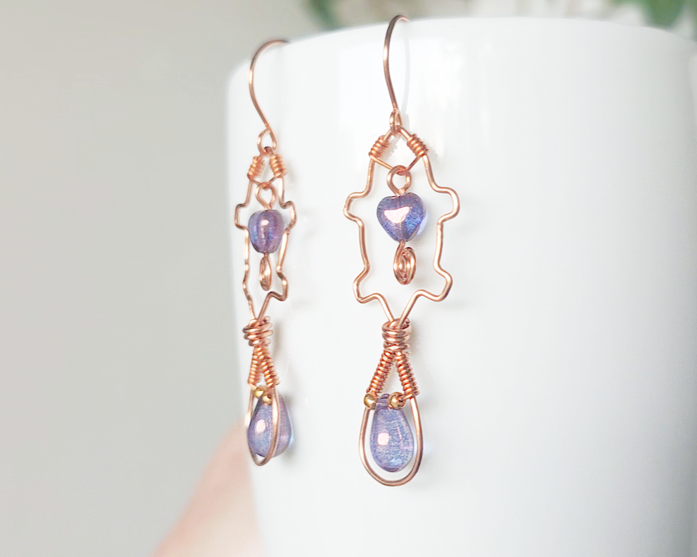 A pair of wire wrapped copper beaver earrings with mauve purple hearts and tear drop stones on french hooks on wood 
