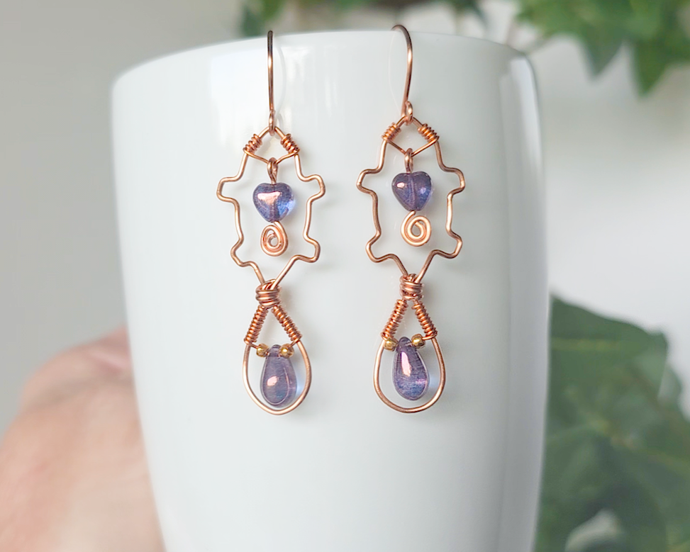A pair of wire wrapped copper beaver earrings with mauve purple hearts and tear drop stones on white vase 
