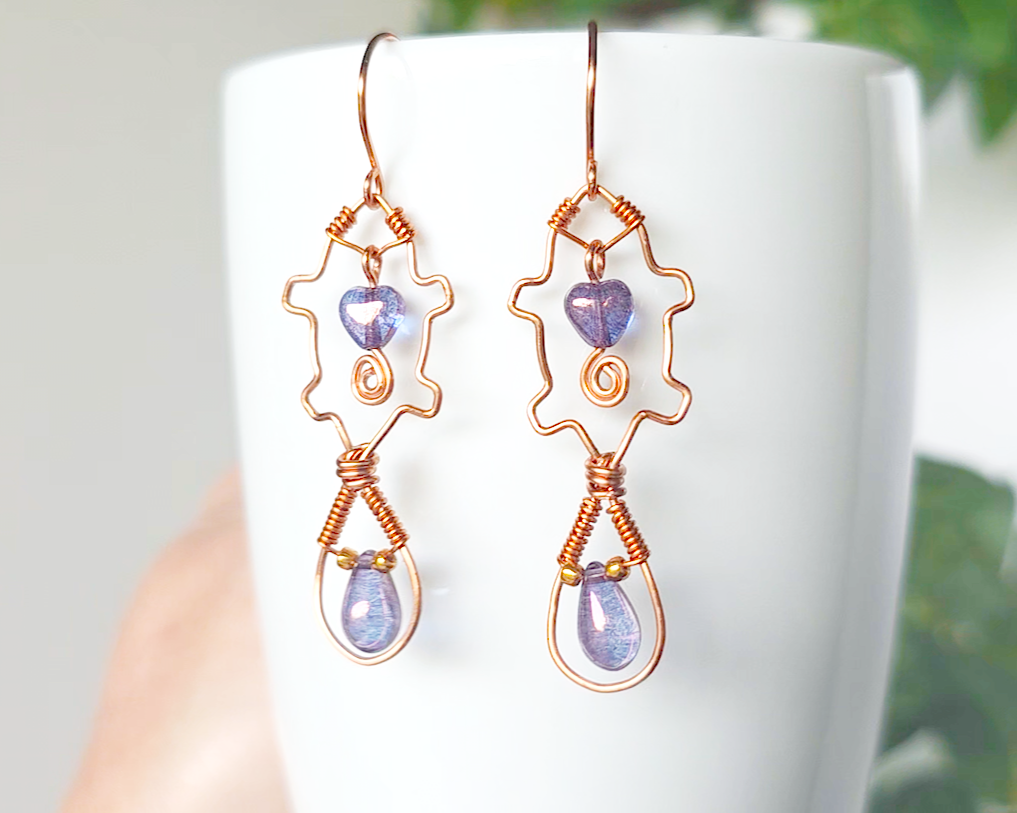 A pair of wire wrapped copper beaver earrings with mauve purple hearts and tear drop stones on french hooks on wood 