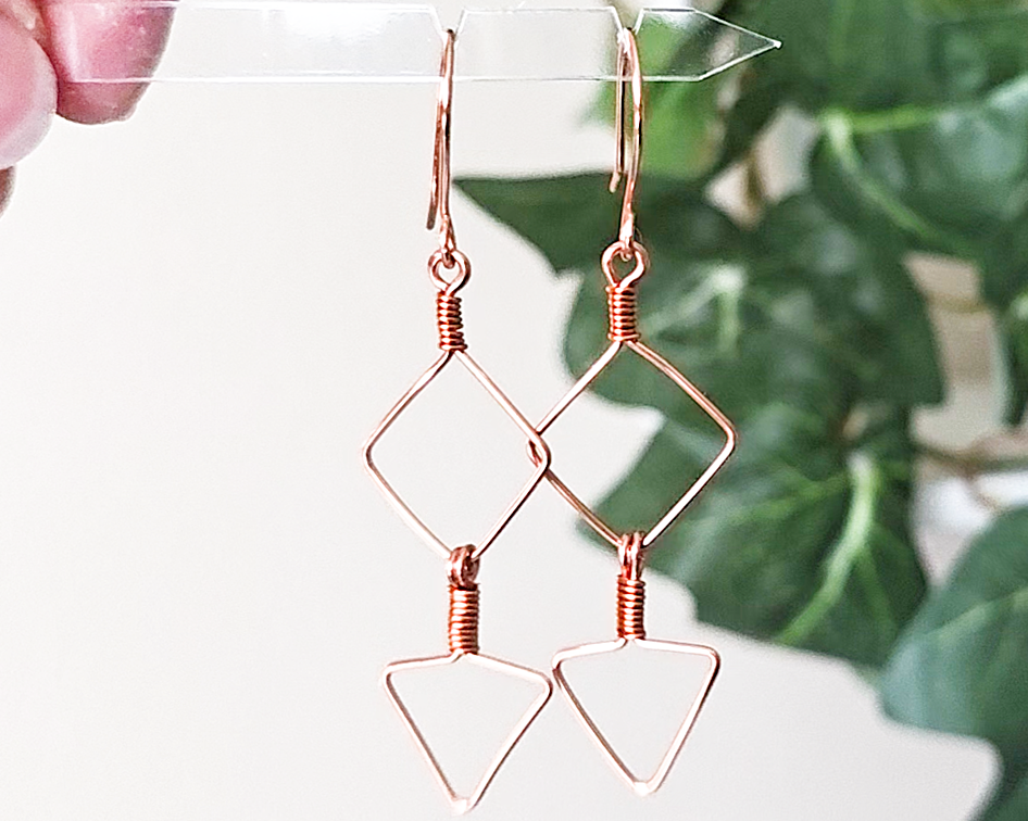 Long Geometric Copper Earrings with Square and Upside down triangles on french earring hooks with green plants in background 