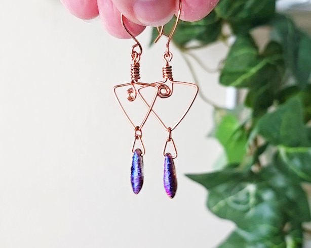 Long Copper Earrings with Celtic Coils in upside down triangle and dagger shaped Iridescent Purple Blue pendants dangling on the base with plants in the background 
