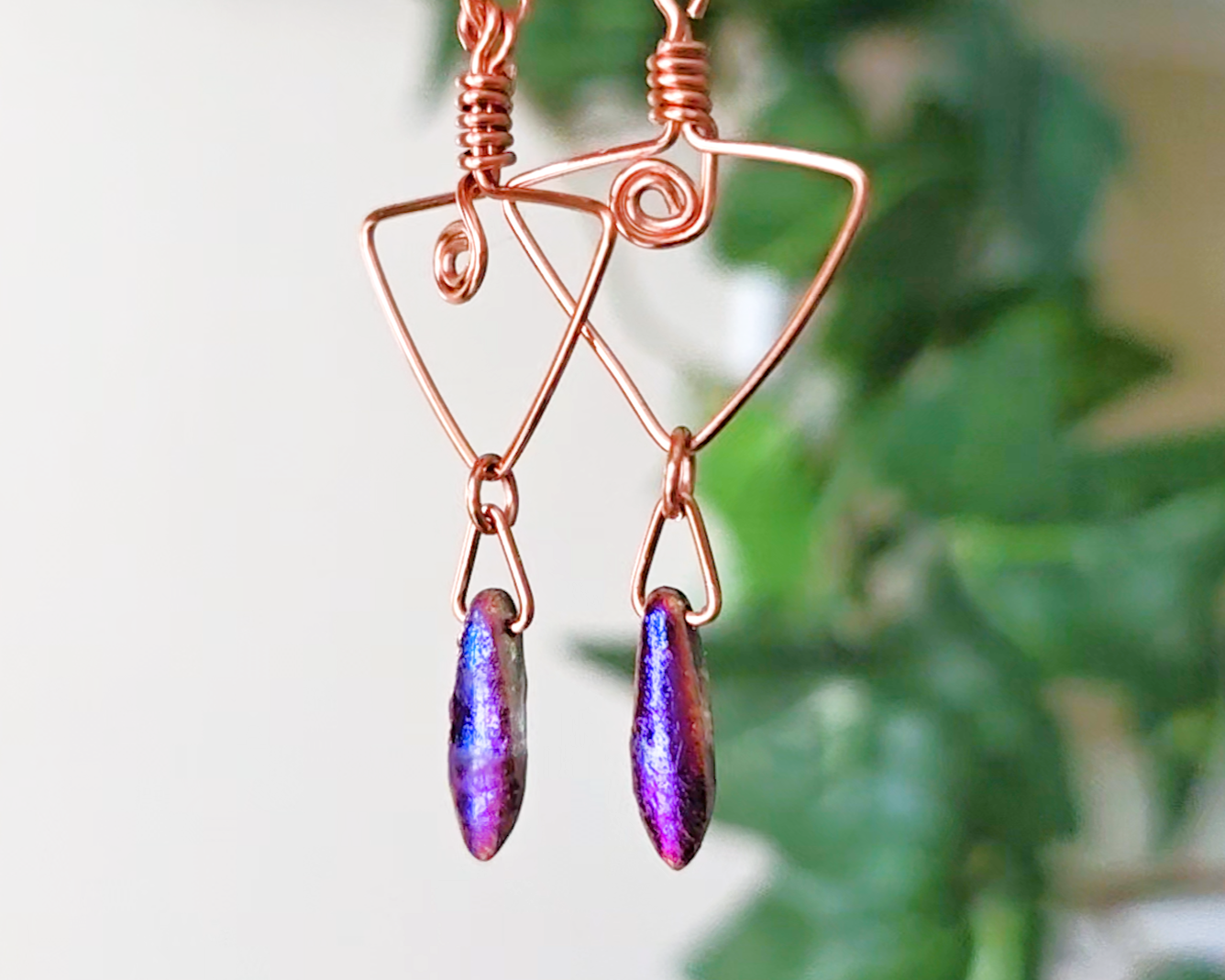 Long Copper Earrings with Celtic Coils in upside down triangle and dagger shaped Metallic Purple Blue pendants dangling on the base with plants in the background 