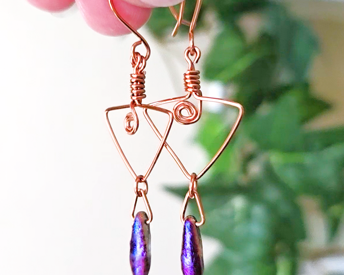 Long Copper Earrings with Celtic Coils in upside down triangle and dagger shaped Metallic Purple Blue pendants dangling on the base with plants in the background 