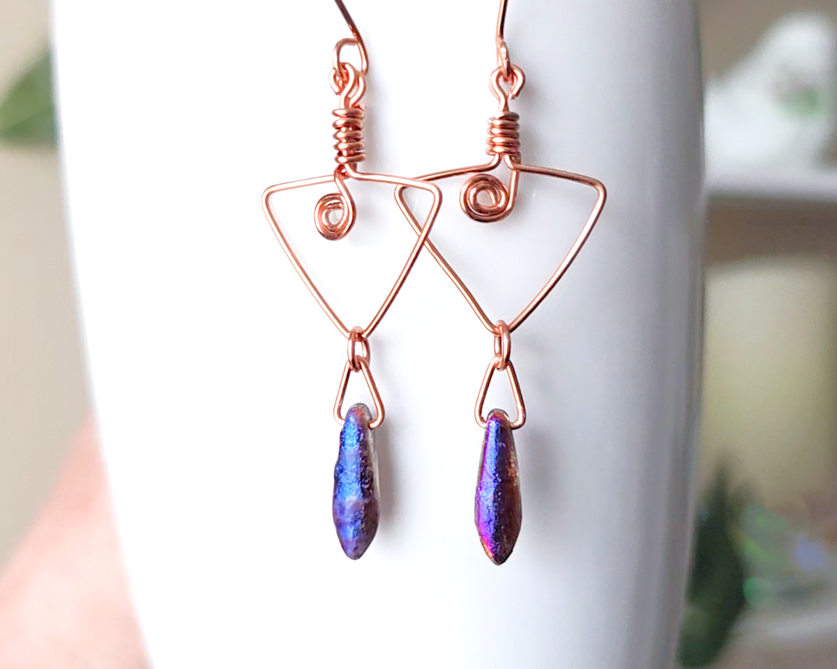 Long Copper Earrings with Celtic Coils in upside down triangle and dagger shaped Metallic Purple Blue pendants dangling on the base displayed on white background 