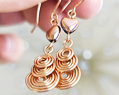 A pair of handmade copper dangle earrings with three coil pendants and gold hearts with french earrings hooks in hand 