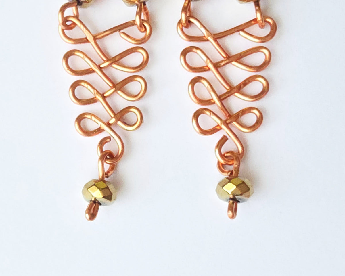  A pair of long handmade copper dangle earrings with a swirling Celtic river design featuring faceted red agates and sparkly faceted gold plated hematite stones on french earrings hooks on white background  