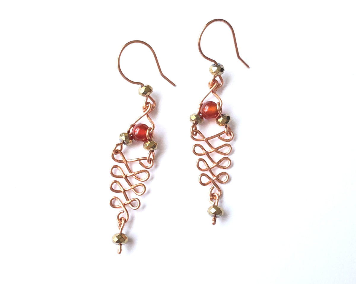  A pair of long handmade copper dangle earrings with a swirling Celtic river design featuring faceted red agates and sparkly faceted gold plated hematite stones on french earrings hooks on white background  