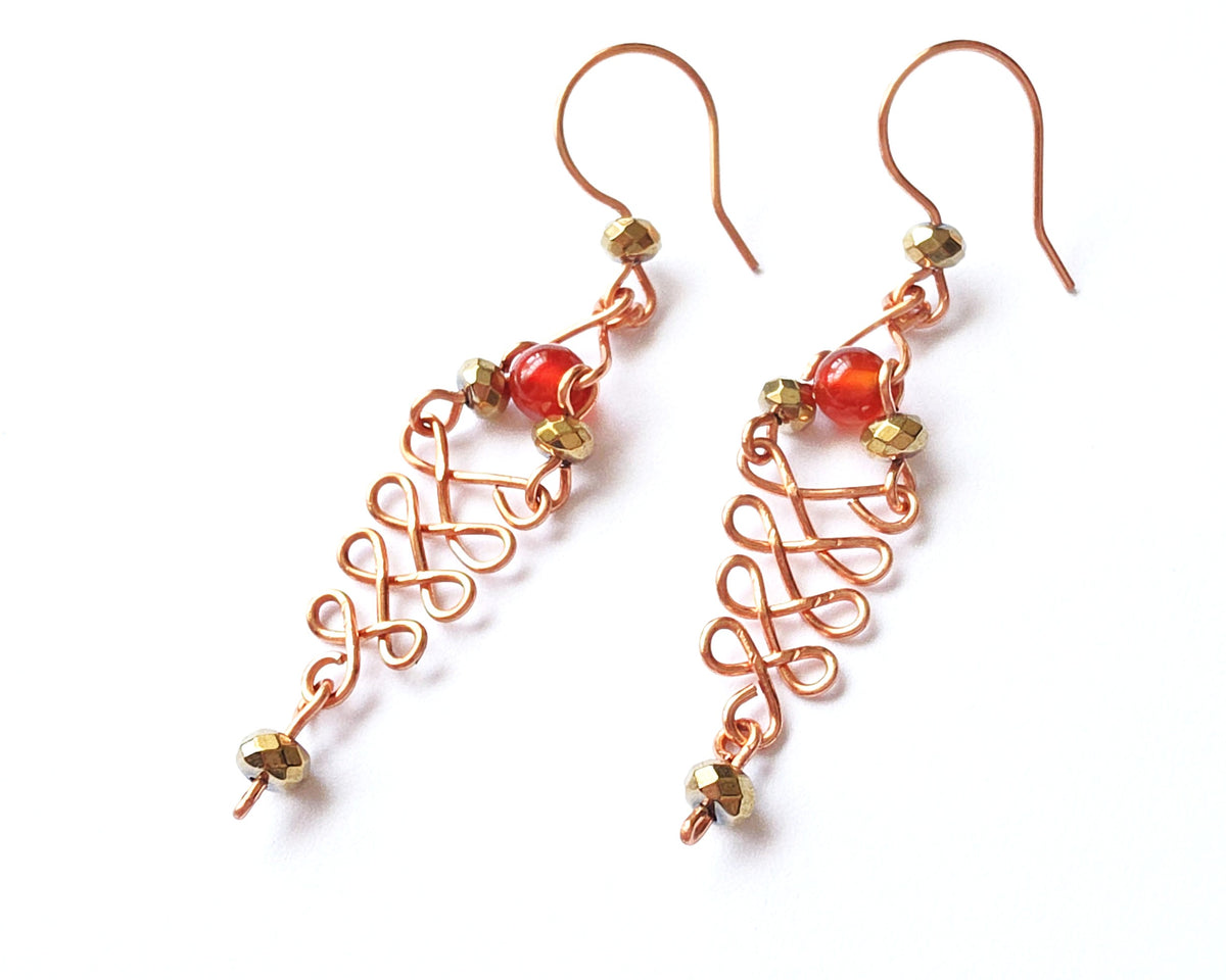  A pair of long handmade copper dangle earrings with a swirling Celtic river design featuring faceted red agates and sparkly faceted gold plated hematite stones on french earrings hooks on white background  