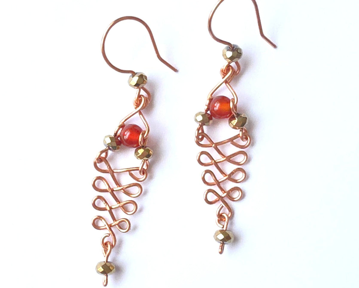  A pair of long handmade copper dangle earrings with a swirling Celtic river design featuring faceted red agates and sparkly faceted gold plated hematite stones on french earrings hooks on white background  