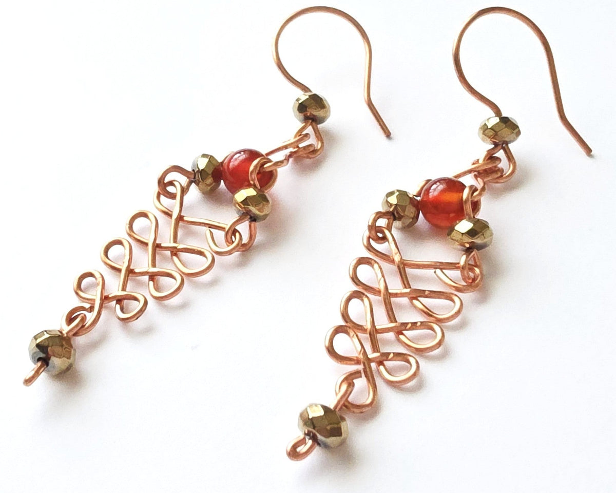  A pair of long handmade copper dangle earrings with a swirling Celtic river design featuring faceted red agates and sparkly faceted gold plated hematite stones on french earrings hooks on white background  
