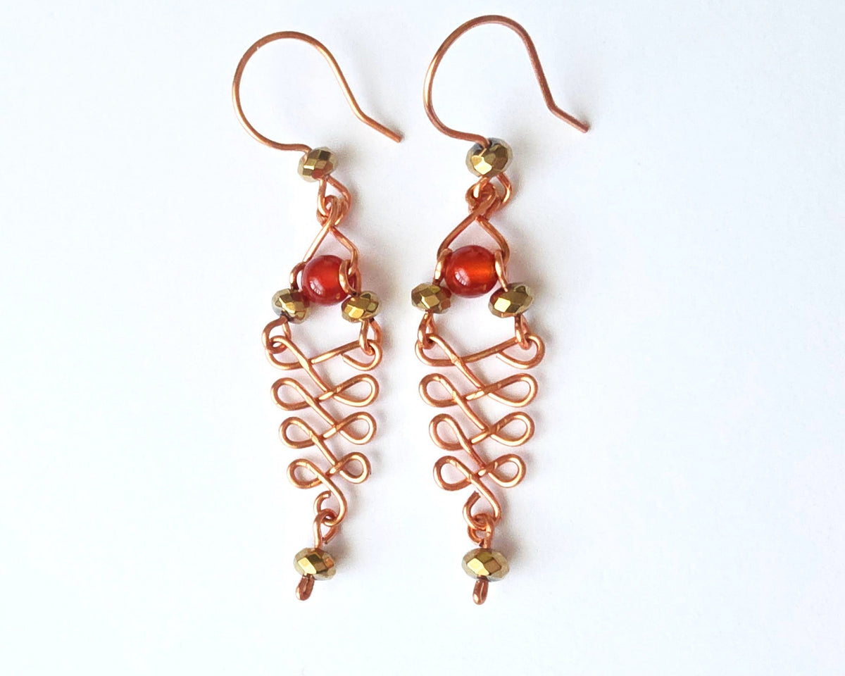 A pair of long handmade copper dangle earrings with a swirling Celtic river design featuring faceted red agates and sparkly faceted gold plated hematite stones on french earrings hooks on white background  