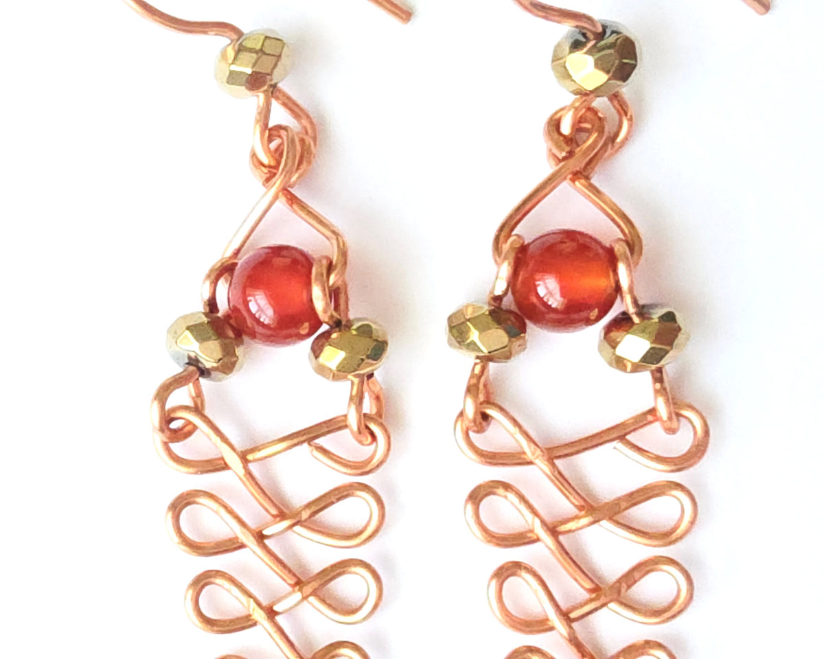  A pair of long handmade copper dangle earrings with a swirling Celtic river design featuring faceted red agates and sparkly faceted gold plated hematite stones on french earrings hooks on white background  