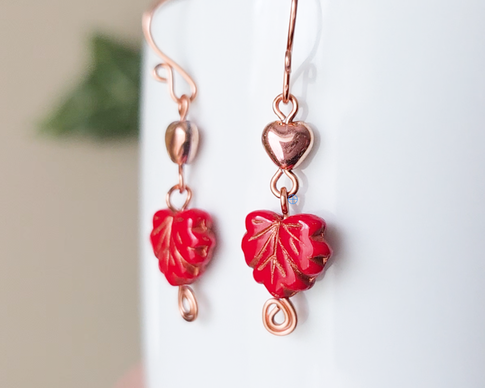 A pair of long handmade copper dangle earrings featuring red glass maple leaf’s, metallic hearts and copper eternity coils on french earring hooks on white background  on white vase green in background 
