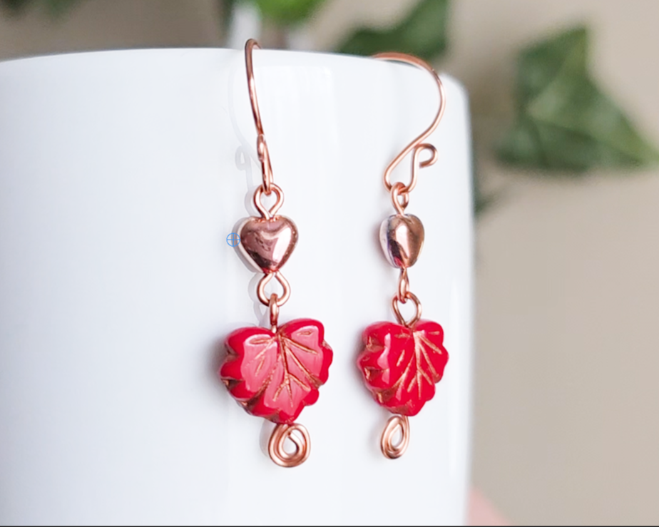 A pair of long handmade copper dangle earrings featuring red glass maple leaf’s, metallic hearts and copper eternity coils on french earring hooks on white background  on white vase green in background 