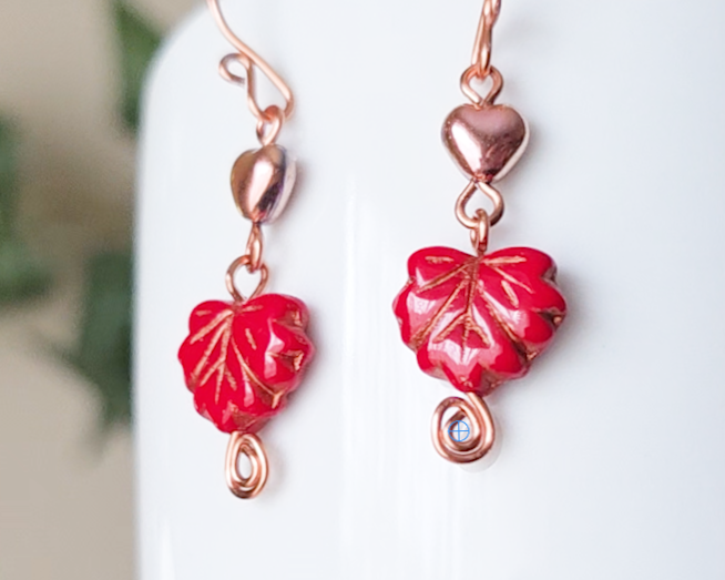A pair of long handmade copper dangle earrings featuring red glass maple leaf’s, metallic hearts and copper eternity coils on french earring hooks on white background  on white vase green in background 
