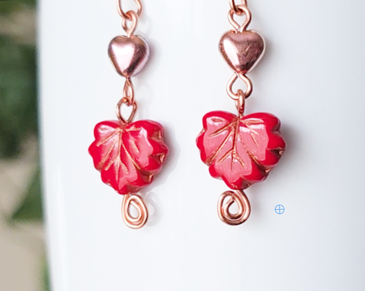 A pair of long handmade copper dangle earrings featuring red glass maple leaf’s, metallic hearts and copper eternity coils on french earring hooks on white background  on white vase green in background 