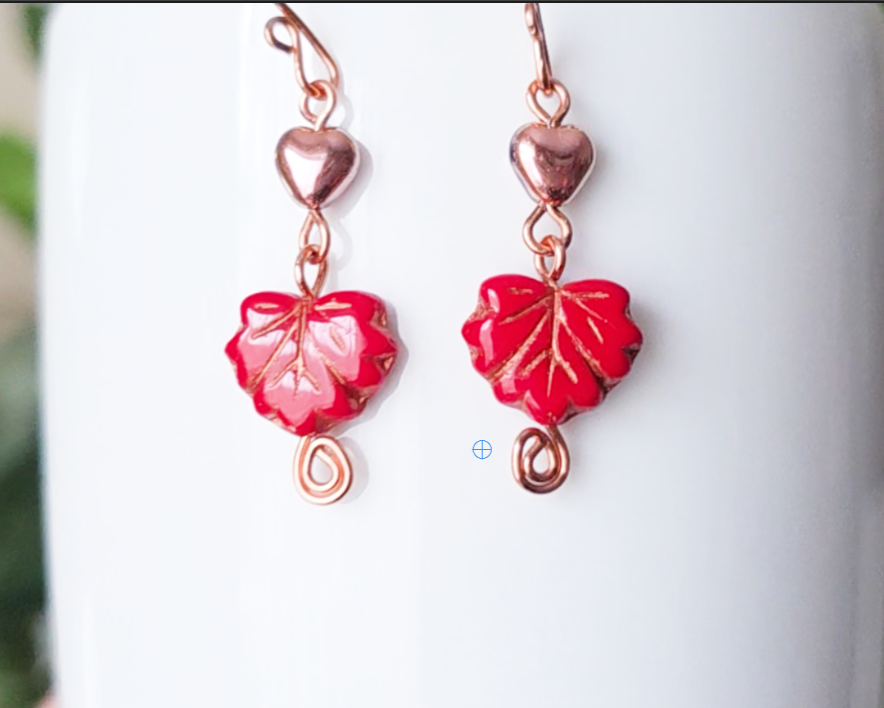 A pair of long handmade copper dangle earrings featuring red glass maple leaf’s, metallic hearts and copper eternity coils on french earring hooks on white background  on white vase green in background 