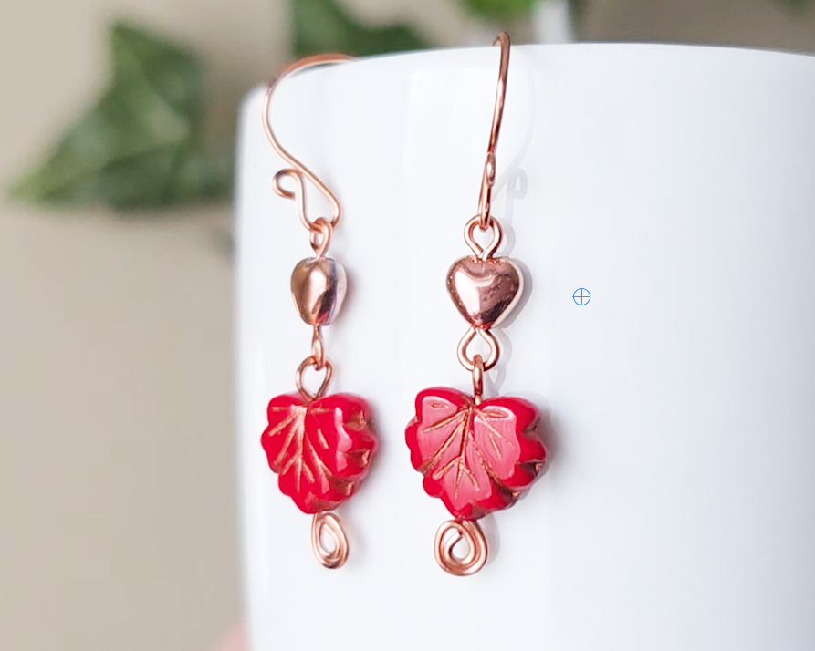 A pair of long handmade copper dangle earrings featuring red glass maple leaf’s, metallic hearts and copper eternity coils on french earring hooks on white background  on white vase green in background 