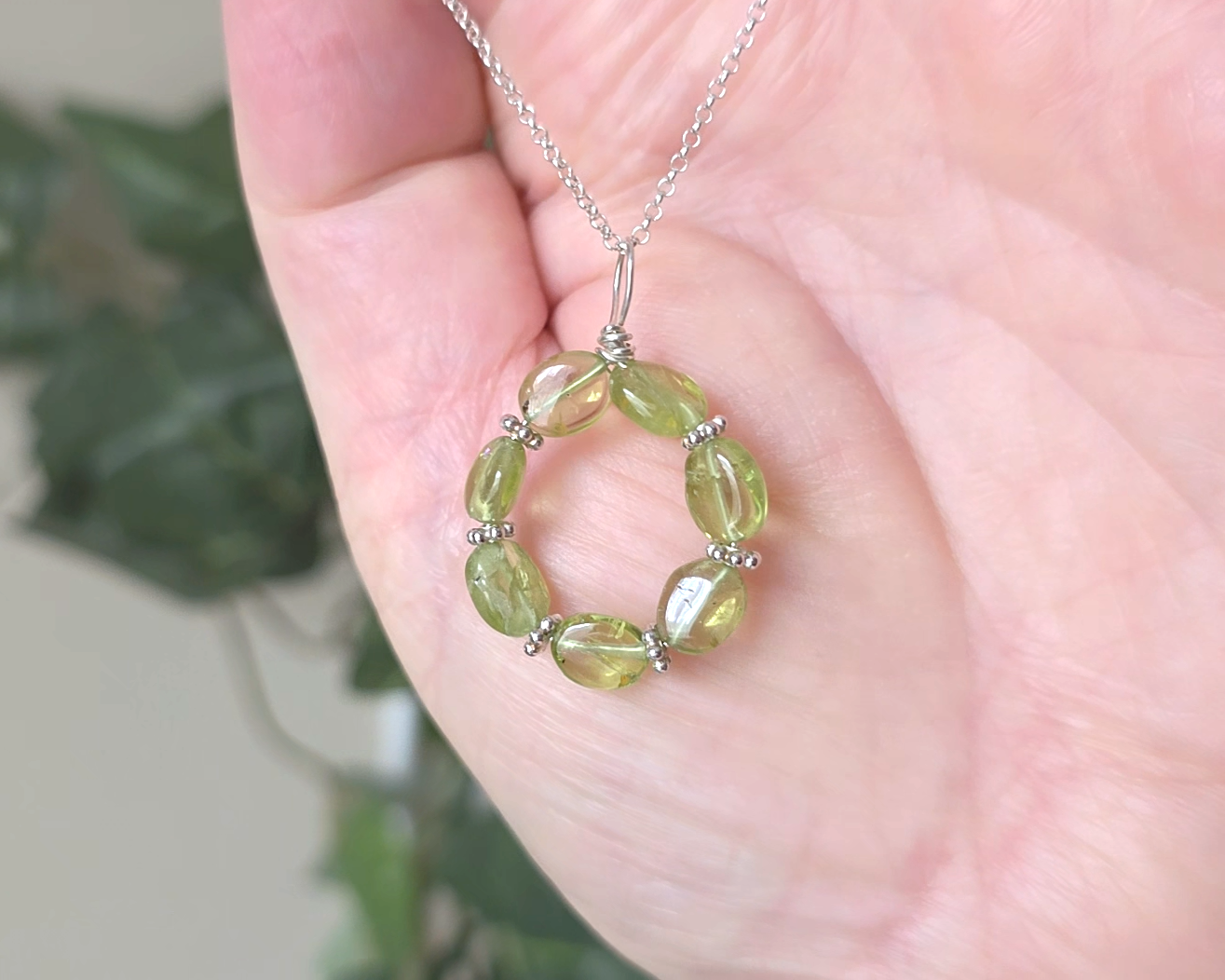 Round Peridot beaded pendant with oval shaped translucent Peridot stones and fancy sterling silver beads in between on a sterling silver rolo chain with plants in hand 