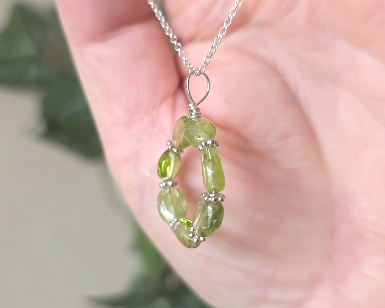 Round Peridot beaded pendant with oval shaped translucent Peridot stones and fancy sterling silver beads in between on a sterling silver rolo chain with plants in hand 