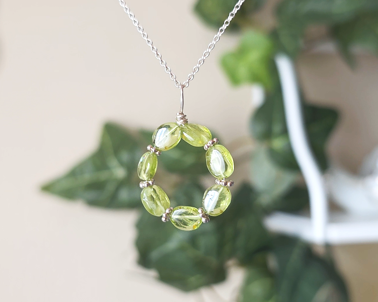 Round Peridot beaded pendant with oval shaped translucent Peridot stones and fancy sterling silver beads in between on a sterling silver rolo chain with plants in the background