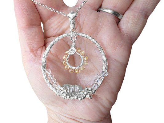 Large Sterling Silver round wire wrapped Sea Glass and Gemstone pendant with aqua sea glass, clear quartz and yellow citrine stones wrapped with silver wire with heart on top and chain, displayed on hand.