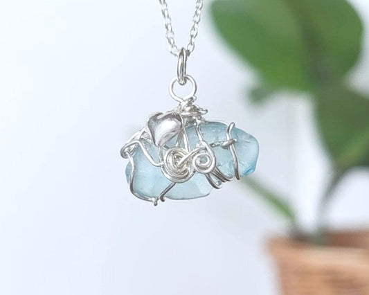 Aqua blue Sea Glass pendant with a Heart & wire wrapped design  in front of a blurred background with plant 