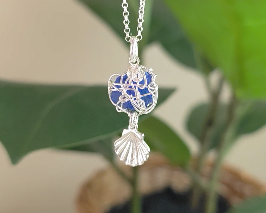 Silver Wire Wrapped Sapphire cobalt blue sea glass pendant with dangling shell on rolo chain with green plants in the background