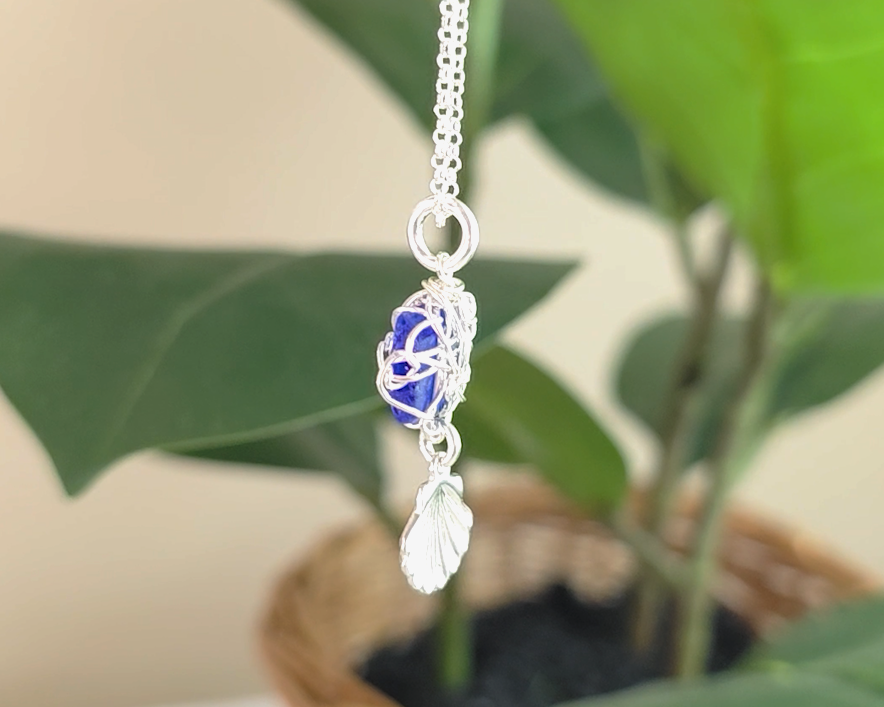 Silver Wire Wrapped Sapphire cobalt blue sea glass pendant with dangling shell on rolo chain with green plants in the background