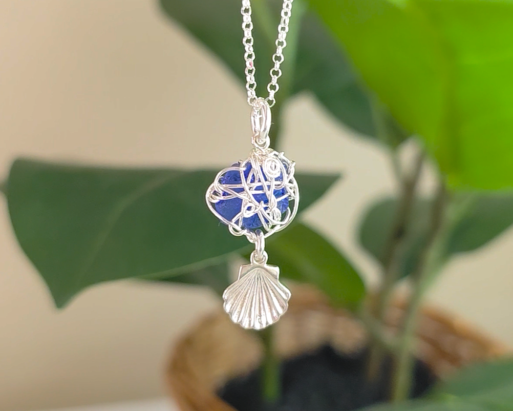 Silver Wire Wrapped Sapphire cobalt blue sea glass pendant with dangling shell on rolo chain with green plants in the background